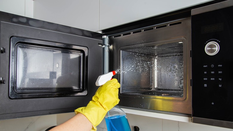 A gloved hand sprays cleaner into a stainless steel microwave