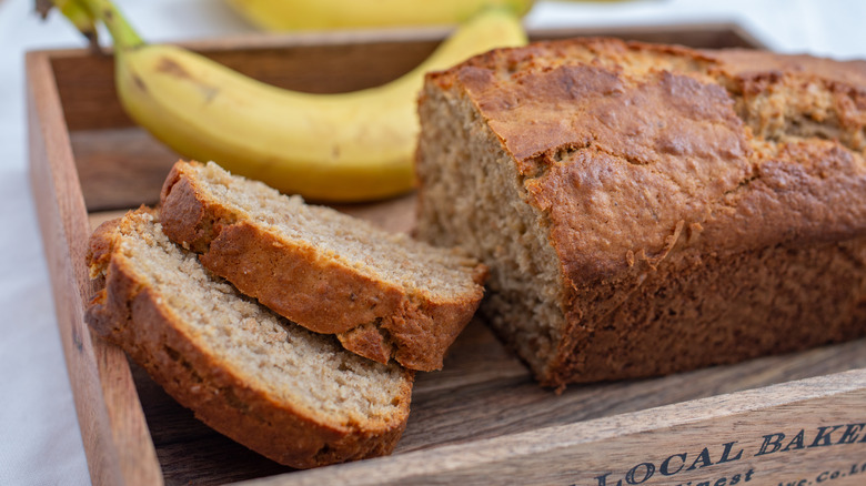 A loaf of banana bread with two sliced pieces sits in a wooden tray, with a banana in the background