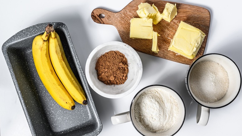 Banana bread-making components sit on a white surface, including a loaf pan holding two unpeeled bananas, dry ingredients in three separate dishes, and butter sitting on a wooden cutting board