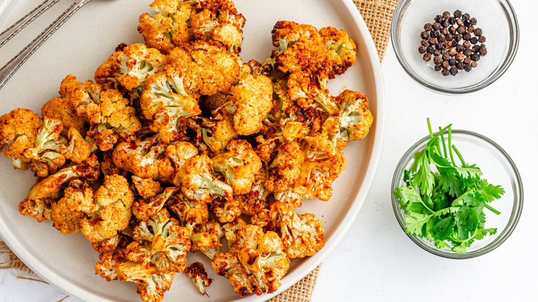 Golden air fried cauliflower on a white place with two forks, on a white surface with two small glass bowls with cilantro and peppercorns