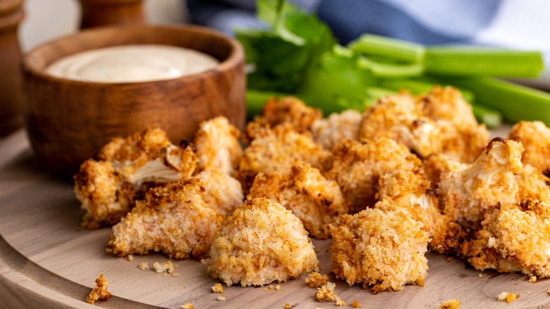 Air fried cauliflower on a wooden tray with a wooden cup of a creamy sauce and celery sticks in the background