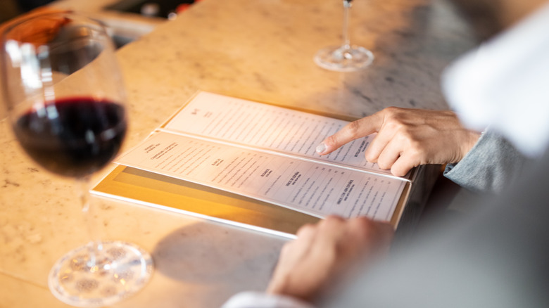 Person pointing at menu card with red wine in foreground