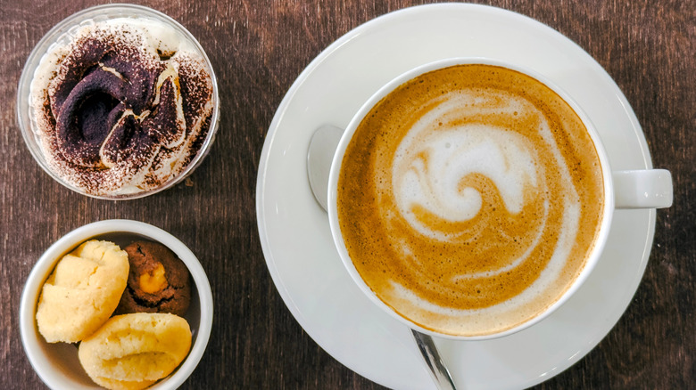 A capuccino in a white coffee mug sits on a wood table with a side of biscotti