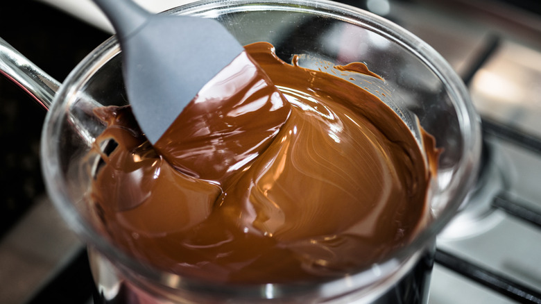 Melted chocolate in a glass bowl over a pot on a stove with a large gray plastic spoon stirring it