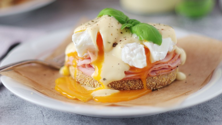 Eggs benedict on English muffin with ham and hollandaise sauce on a white plate
