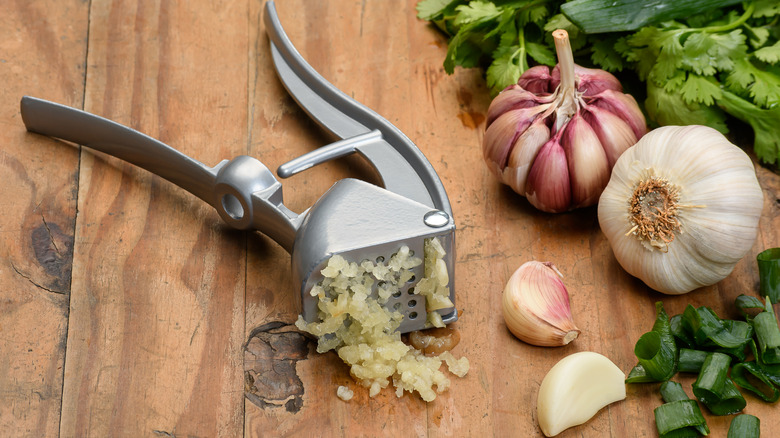 Garlic press with garlic cloves, cilantro, and chopped green onions on wood table