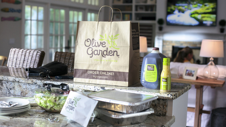 A batch of Olive Garden delivery sits in someone's kitchen, complete with jug of drink, bottle of salad dressing, two tins of food, a bag of breadsticks, and a container of salad