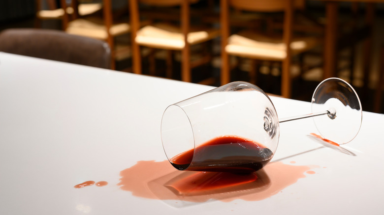 A glass of spilled red wine sits on its side beneath a red wine stain on a white tablecloth