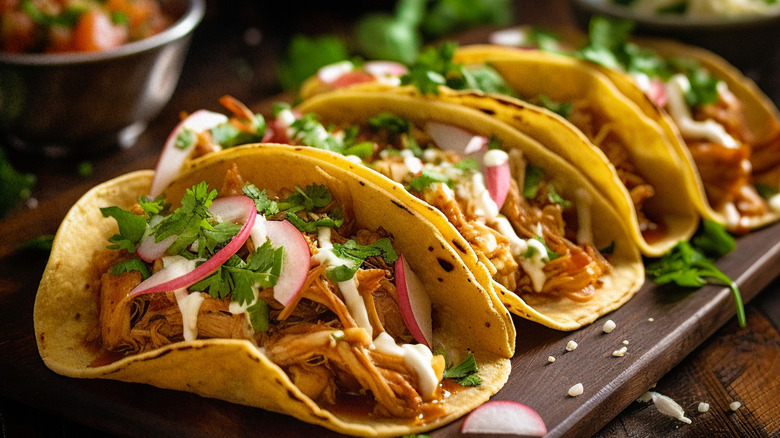 Four shredded meat tacos in corn tortillas sit on a wooden plank