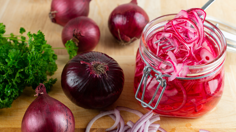 Pickled red onions are lifted by fork from a glass jar, sitting next to whole and chopped red onions on a wooden counter