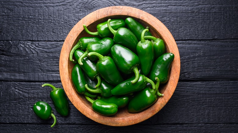 Green jalapeños in a wooden bowl on a black surface with some loose jalapeños on the counter