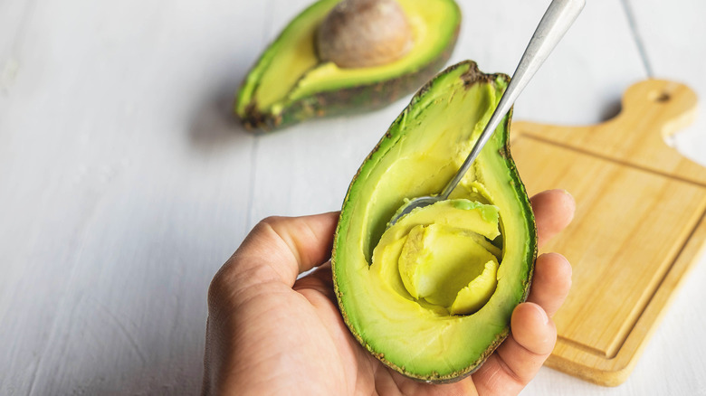 A hand holds half an avocado while a spoon is scooping out the flesh.