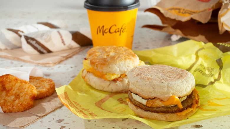 McDonald's breakfast items sit on a countertop, including a Sausage McMuffin with Egg, a Bacon, Egg & Cheese Biscuit, hash browns, and a McCafe coffee