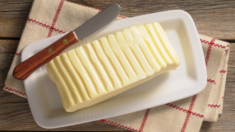 Stick of scored butter on white rectangular dish next to butter knife