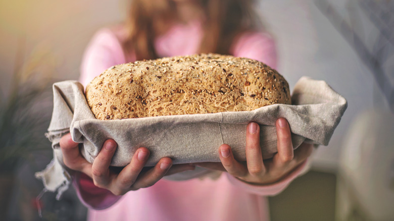 A girl holds up a loaf of homemade bread covered in seeds and wrapped in a tea towel