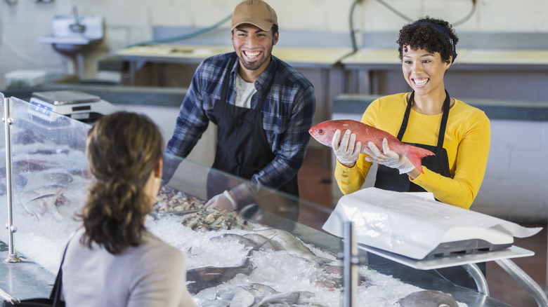 A customer chats with two workers behind a seafood counter, one holding up a red fish