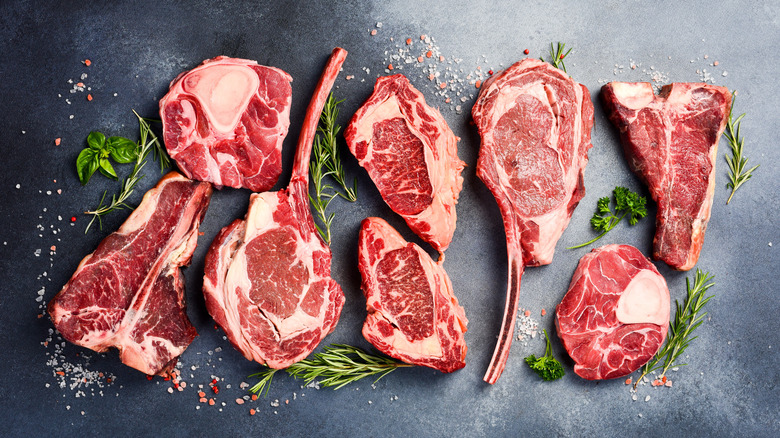 A variety of steaks are sitting on a counter with herbs and seasonings.