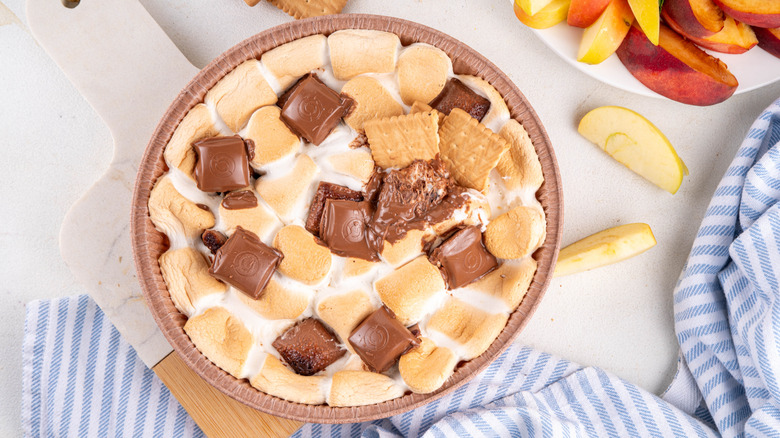 A finished graham cracker s'mores dip sits on a counter beside a selection of fruit slices.