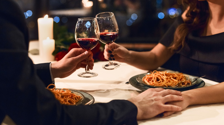 A couple holds hands while toasting with red wine and pasta on a candlelit table