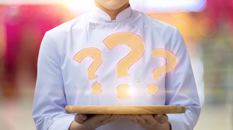 A person in a chef's coat holds a wooden tray with question marks hovering above it.