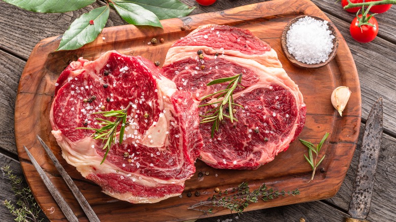 Two raw steaks with salt, peppercorns, and rosemary on a wooden cutting board