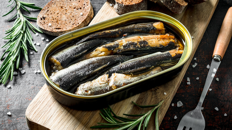 tin of sardines on a breadboard with bread and sprig of rosemary