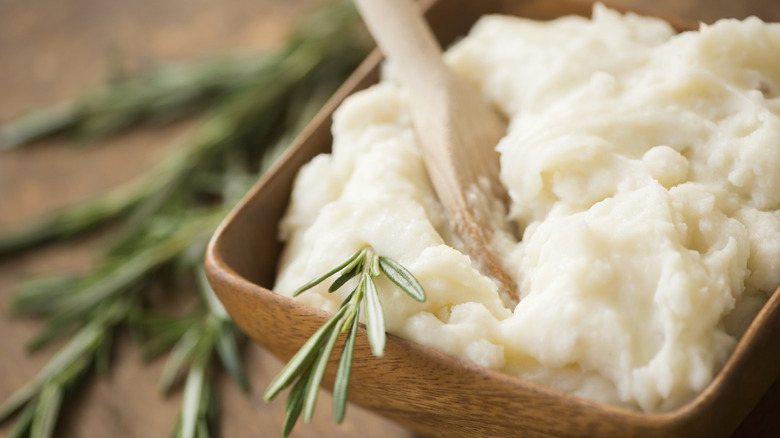 Mashed potatoes in a wooden bowl with a wooden spoon and a spring of herbs