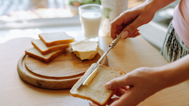 A person spreads butter on bread with bread slices and butter on a wooden tray and a glass of milk in the background of a brightly lit kitchen