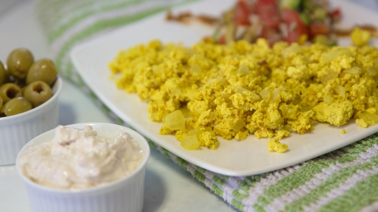 Bright-yellow tofu scramble on a white plate next to green olives and cottage cheese