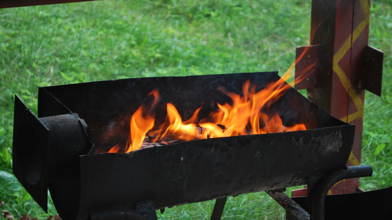 An open grill with flames is burning near a grassy background.