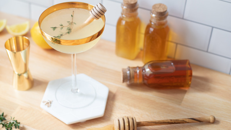 Pale white drink in a gold-rimmed cocktail glass with a sprig of thyme and a mini honey dipper as garnish