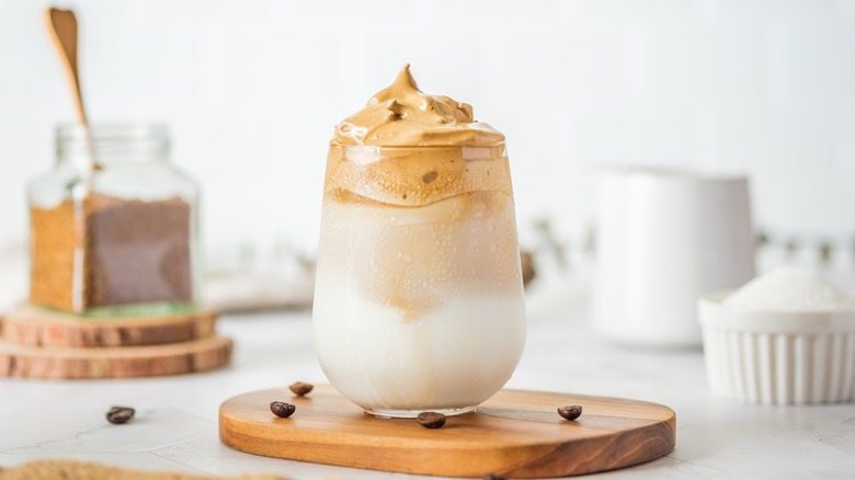 Dalgona coffee layered with whipped foam, on wooden tray. Coffee beans spread on table, with ingredients blurred in the background.