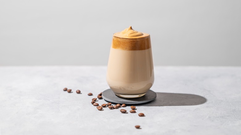 Dalgona coffee showing layers of milk and foam in a clear glass, on top of a coaster. Coffee beans are surrounding the coaster, all on a texturized white background.