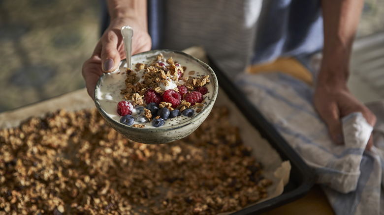 A person holds a bowl of granola and yogurt over a baking tray of fresh granola