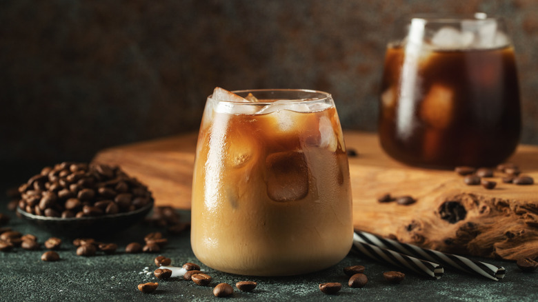 Iced coffee in a short glass with cream or milk. Coffee beans are surrounding the glass with two straws behind. A second glass of black iced coffee sitting on a wooden tray, and blurred behind. All on a dark, texturized background.