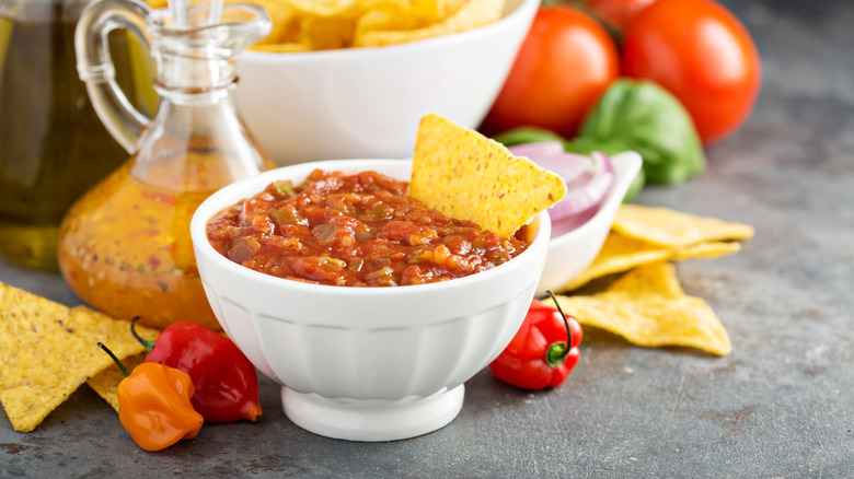 A bowl of homemade salsa with a tortilla chip surrounded by habanero peppers, tomatoes, and basil