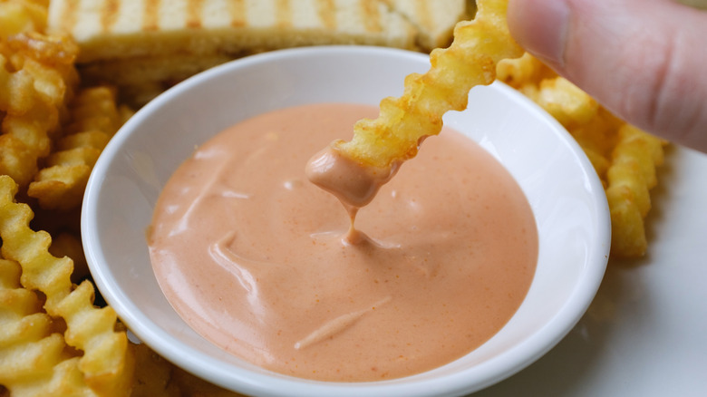 A hand dips a crinkle-cut french fry into a white saucer of fry sauce