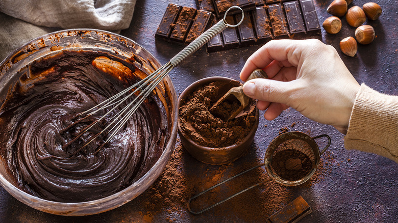 A hand scoops cocoa powder from a ramekin that sits beside a mixing bowl filled with chocolate batter, a sifter, bricks of chocolate, and hazelnuts