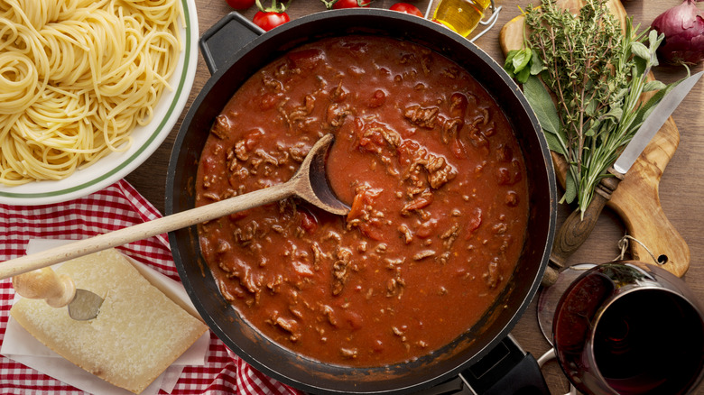 A pan full of bolognese sauce rests next to a bowl of spaghetti, fresh herbs, cheese, and other Italian-inspired accoutrements