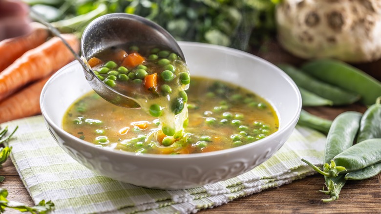 Ladle in bowl of broth-based vegetable soup surrounded by veggies