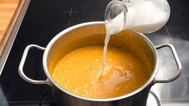 Milk poured from a clear glass pitcher into a pot of soup