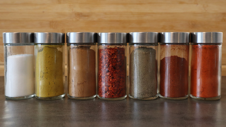 Seven glass spice bottles lined up on a counter, wooden wall behind them