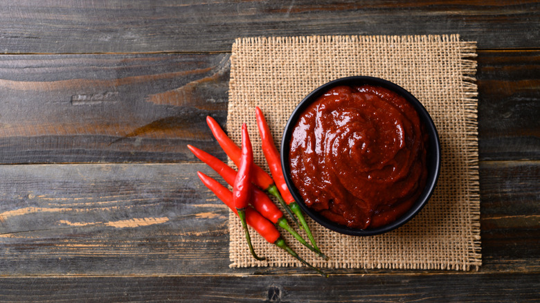 Gochujang in a black bowl on a wooden table next to red chile peppers.