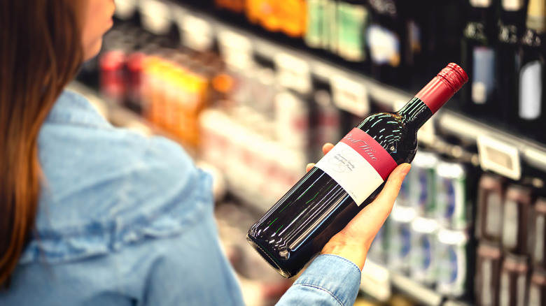 A woman in a blue shirt looks at a bottle of wine while shopping in a grocery store