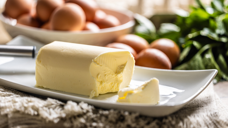 A stick of fresh butter sits on a plate in front of some brown eggs and fresh greens