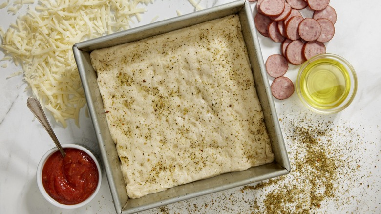 A raw Detroit-style pizza dough is in a square pan, sprinkled with herbs, with ramekins of sauce and oil and piles of sliced meat and grated cheese on the counter adjacent