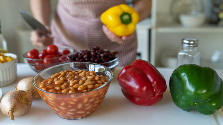 Onions, bowl of baked beans, and red and green pepper on a kitchen counter with person in background holding yellow pepper and knife