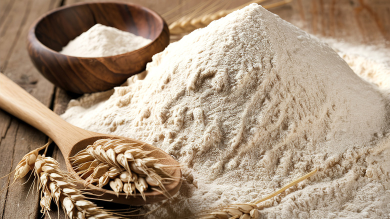 A pile of flour sits on a wooden surface near a small wooden bowl containing flour and a wooden spoon holding grains of wheat