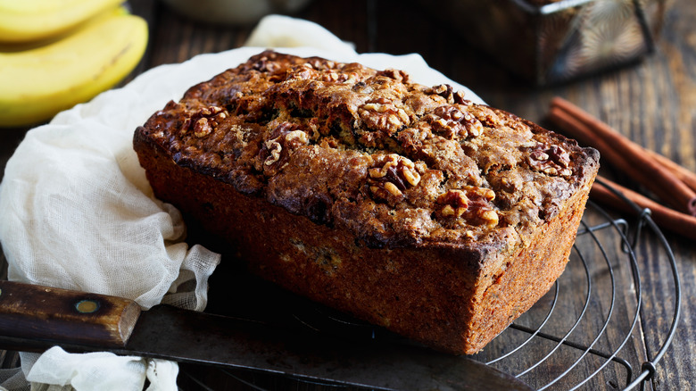 Banana bread sitting on a wire rack with bananas in the background and a knife in the foreground