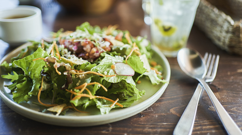 A plate of green salad sits on a table next to a fork and spoon, with beverages sitting adjacent in the background